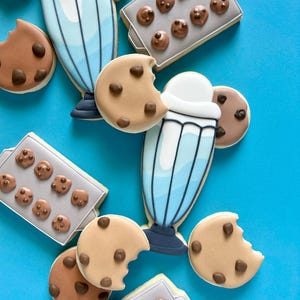 May include: An assortment of decorated sugar cookies on a bright blue surface. The cookies include chocolate chip cookies, some with a bite taken out, and milkshake-shaped cookies. There are also cookies shaped like baking trays with mini chocolate chip cookies.
