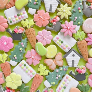 May include: A collection of spring-themed sugar cookies decorated with various designs, including flowers, butterflies, carrots, watering cans, and houses. The cookies are arranged on a green background.