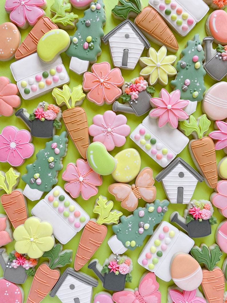 May include: A collection of spring-themed sugar cookies decorated with various designs, including flowers, butterflies, carrots, watering cans, and houses. The cookies are arranged on a green background.
