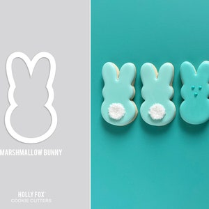 May include: A set of five bunny-shaped sugar cookies decorated with pastel blue and pink icing. The cookies have white sprinkles on the back and some have small blue sprinkles on the front. The cookies are arranged in a row on a turquoise background. The image also includes a white outline of a bunny cookie cutter.