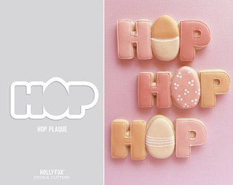 May include: Three sets of pink and orange cookies decorated with icing in the shape of the word "HOP" with an Easter egg in the center of each set. The Easter eggs are decorated with white icing.