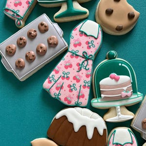 May include: Assortment of decorated sugar cookies in various shapes, including a dress, oven mitt, cake, and chocolate chip cookies. The cookies are decorated with pink, green, white, and brown icing, and arranged on a teal background.