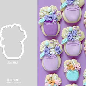 May include: A set of nine cookie cutters shaped like egg vases with floral designs. The cookie cutters are made of white plastic and are arranged on a purple background. The text "EGG VASE" and "HOLLY FOX COOKIE CUTTERS" is visible on the image.