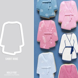 May include: A set of six cookie cutters shaped like short robes. The cookie cutters are made of white metal and are arranged in a row on a blue background. The cookie cutters are labeled "Short Robe" and "Holly Fox Cookie Cutters".