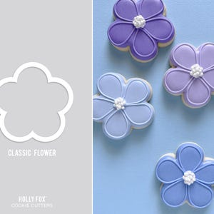 May include: A white flower-shaped cookie cutter next to several flower-shaped cookies. The cookies are decorated with shades of purple and blue icing and white accents. The text "CLASSIC FLOWER" and "HOLLY FOX COOKIE CUTTERS" are also visible.