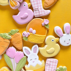 May include: A collection of Easter-themed sugar cookies decorated with various designs, including a bunny, a duck, a basket of eggs, carrots, tulips, and a butterfly. The cookies are arranged on a yellow background.