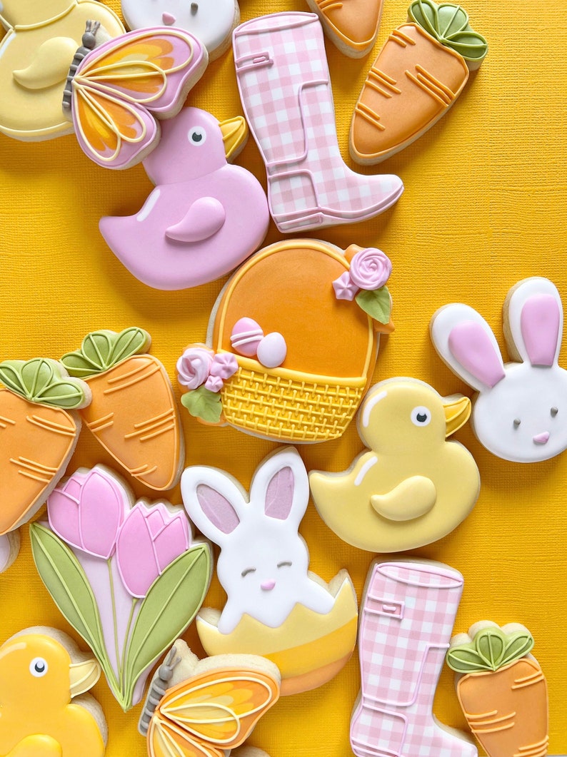 May include: A collection of Easter-themed sugar cookies decorated with various designs, including a bunny, a duck, a basket of eggs, carrots, tulips, and a butterfly. The cookies are arranged on a yellow background.