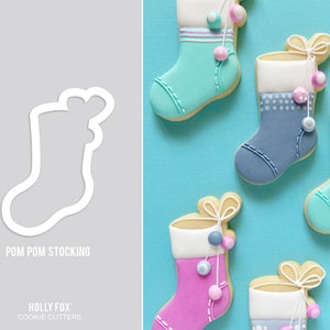 May include: A set of five cookie cutters shaped like Christmas stockings with pom-poms. The cutters are made of white plastic and are arranged on a blue background. The text "POM POM STOCKING" is printed below the cutters. The text "HOLLY FOX COOKIE CUTTERS" is printed below the text "POM POM STOCKING".