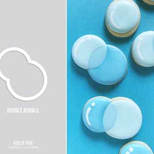 May include: Double bubble cookie cutter, a white outline of two overlapping circles on a gray background.  The cutter is shown on a blue background with several cookies decorated with blue icing to look like bubbles.