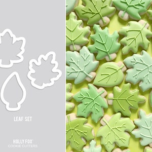 May include: A set of three white leaf-shaped cookie cutters next to leaf-shaped cookies with green frosting. The cookies have detailed leaf vein designs. The text "LEAF SET" and "HOLLY FOX COOKIE CUTTERS" are visible.