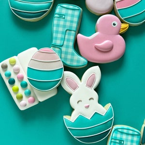 May include: Assortment of Easter-themed sugar cookies decorated with pastel colors and various designs, including a bunny in an egg, a duck, a boot, and an egg with a pink and white stripe.