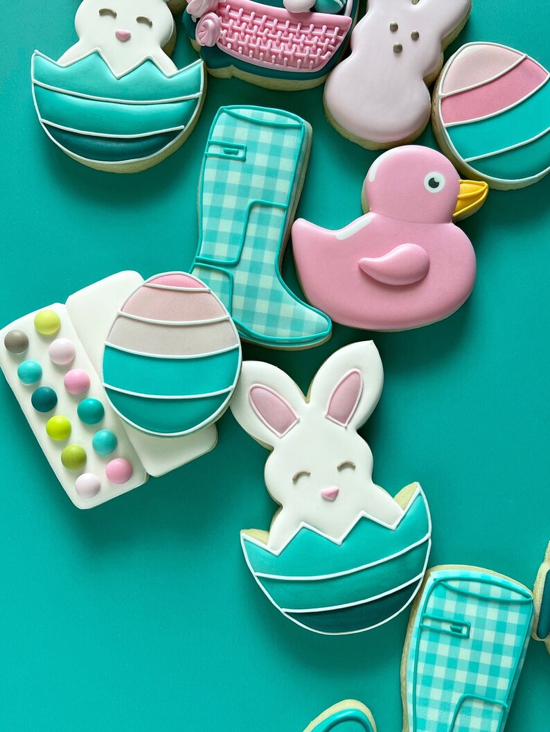 May include: Assortment of Easter-themed sugar cookies decorated with pastel colors and various designs, including a bunny in an egg, a duck, a boot, and an egg with a pink and white stripe.