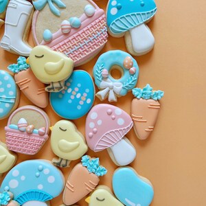 May include: A collection of Easter-themed biscuits. The biscuits are decorated in pastel shades and feature designs such as mushrooms, carrots, chicks, Easter eggs, and baskets. They are arranged on an orange background.