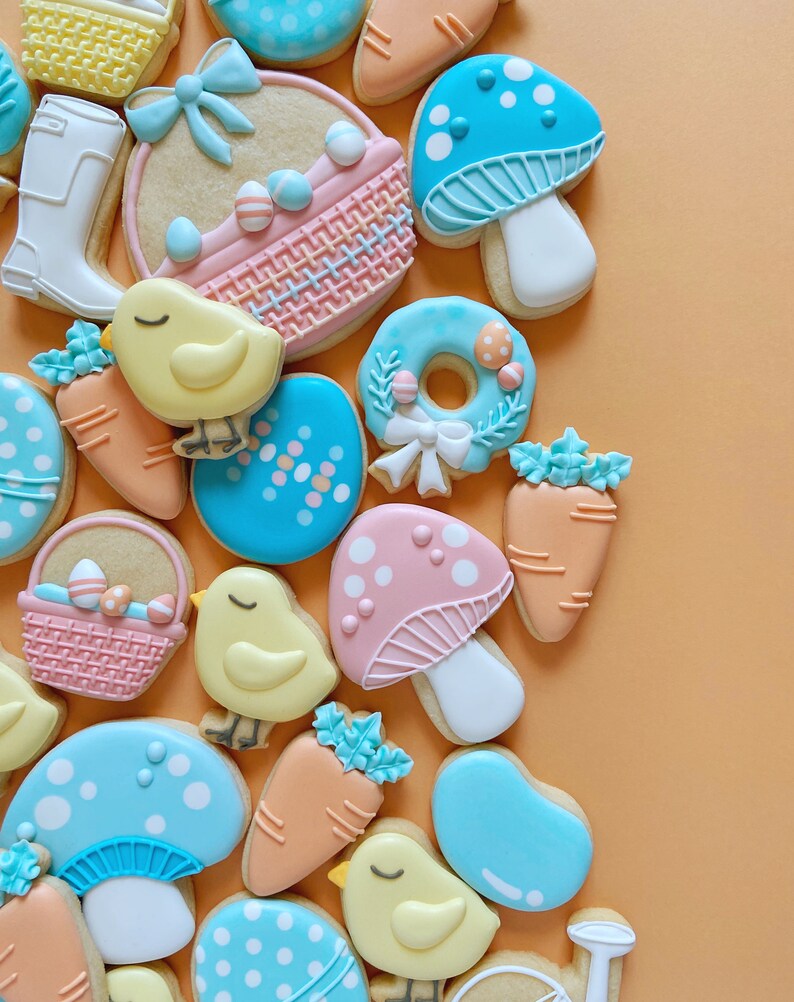 May include: A collection of Easter-themed biscuits. The biscuits are decorated in pastel shades and feature designs such as mushrooms, carrots, chicks, Easter eggs, and baskets. They are arranged on an orange background.
