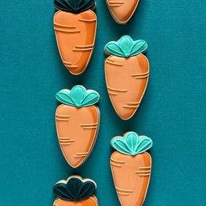 May include: Six carrot-shaped biscuits with orange icing and green and dark green leaf details. The biscuits are arranged vertically on a teal background. Each biscuit has horizontal lines to represent the texture of a carrot.
