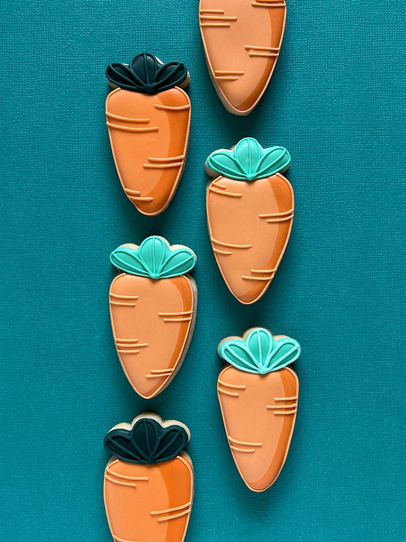 May include: Six carrot-shaped biscuits with orange icing and green and dark green leaf details. The biscuits are arranged vertically on a teal background. Each biscuit has horizontal lines to represent the texture of a carrot.