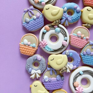 Op de afbeelding: Een verzameling paas-suikerkoekjes versierd met pastelkleuren en verschillende ontwerpen, waaronder manden, kuikens, kransen en donuts.