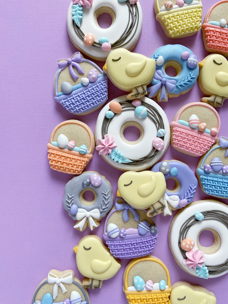 Op de afbeelding: Een verzameling paas-suikerkoekjes versierd met pastelkleuren en verschillende ontwerpen, waaronder manden, kuikens, kransen en donuts.
