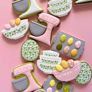 May include: Assortment of Easter-themed cookies on a pink background. The cookies include stand mixers, mixing bowls, and eggs. The cookies are decorated with pastel colors, floral patterns, and dots. The cookies are arranged in a visually appealing manner.