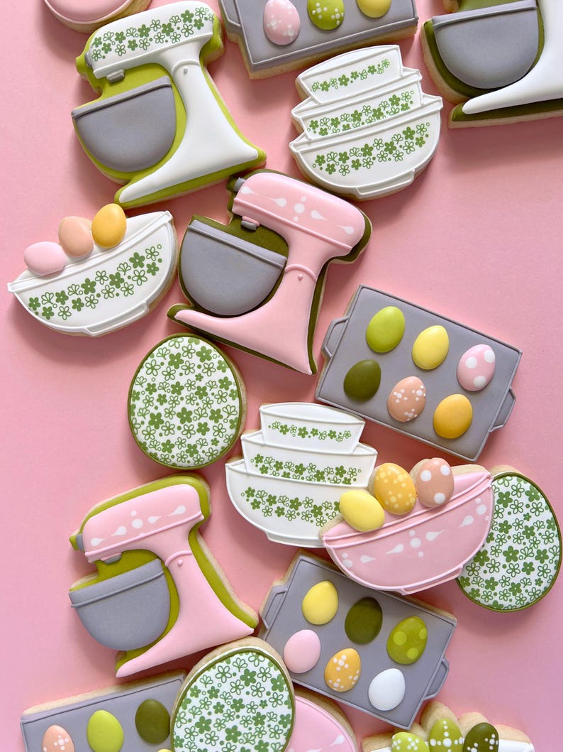 May include: Assortment of Easter-themed cookies on a pink background. The cookies include stand mixers, mixing bowls, and eggs. The cookies are decorated with pastel colors, floral patterns, and dots. The cookies are arranged in a visually appealing manner.