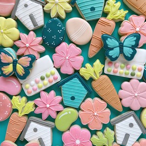 May include: An assortment of colourful sugar cookies decorated with various spring and Easter themes, including butterflies, flowers, carrots, birdhouses and eggs. The cookies are arranged on a turquoise background.