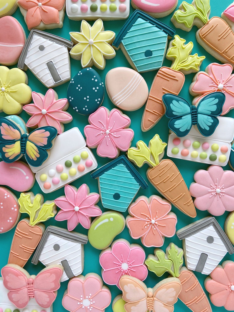 May include: An assortment of colourful sugar cookies decorated with various spring and Easter themes, including butterflies, flowers, carrots, birdhouses and eggs. The cookies are arranged on a turquoise background.