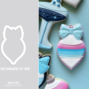 May include: A cookie cutter in the shape of a vintage Christmas ornament with a bow. The cutter is white and is shown against a gray background. The text "Vintage Ornament w/ Bow" is below the cutter. The text "HOLLY FOX COOKIE CUTTERS" is below the text.