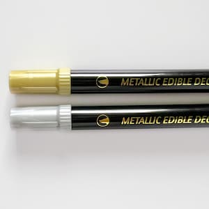 May include: Two black edible decorating markers with gold caps. The top marker has a gold tip and the bottom marker has a silver tip. The markers say "Metallic Edible Decoration" in gold lettering.