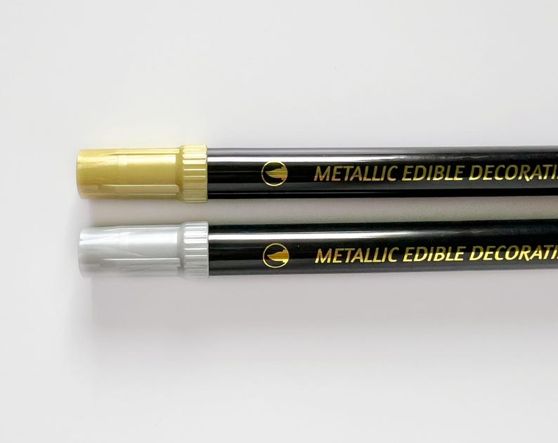 May include: Two black edible decorating markers with gold caps. The top marker has a gold tip and the bottom marker has a silver tip. The markers say "Metallic Edible Decoration" in gold lettering.