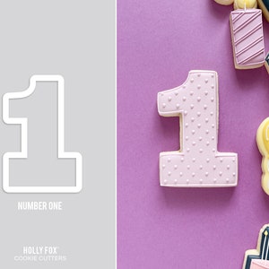 May include: A white cookie cutter in the shape of the number one, with the text "NUMBER ONE" below it. A frosted, pink cookie in the shape of the number one is decorated with small white hearts. Other decorated cookies are also visible.