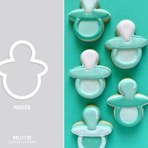 May include: Cookies iced to resemble baby pacifiers, in shades of teal and white, arranged on a teal surface. A cookie cutter outline of a pacifier is on a grey background with the word "PACIFIER".