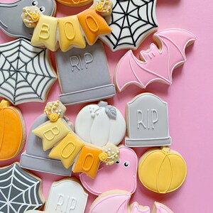 May include: A collection of Halloween-themed sugar cookies decorated with various designs, including ghosts, bats, pumpkins, spiderwebs, and tombstones. The cookies are arranged on a pink background.