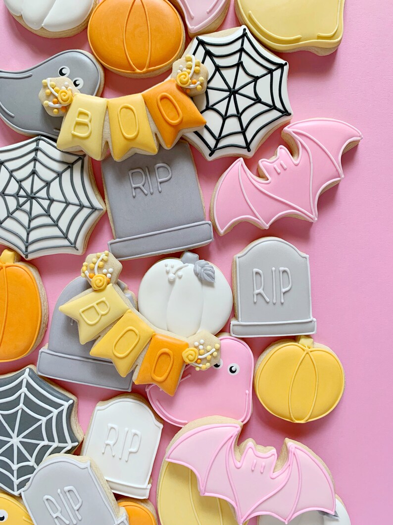 May include: A collection of Halloween-themed sugar cookies decorated with various designs, including ghosts, bats, pumpkins, spiderwebs, and tombstones. The cookies are arranged on a pink background.