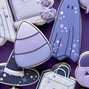 Puede incluir: Galletas de az&uacute;car de Halloween de color morado y blanco decoradas con varios dise&ntilde;os, incluyendo un fantasma, una l&aacute;pida con la palabra "RIP", un sombrero de bruja, un ma&iacute;z dulce, una bolsa de compras y una calabaza.