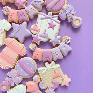 以下が含まれることがあります： ベビーをテーマにしたデコレーションされたシュガー クッキーのコレクション。クッキーはピンク、パープル、ホワイトのアイシングで飾られており、星、ベビーラトル、ベビーモバイル、そして「BABY」と書かれたバナーなどのデザインが施されています。