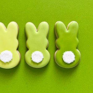 May include: Three green bunny-shaped sugar cookies with white sprinkles on the back. The cookies are arranged in a row on a green background.