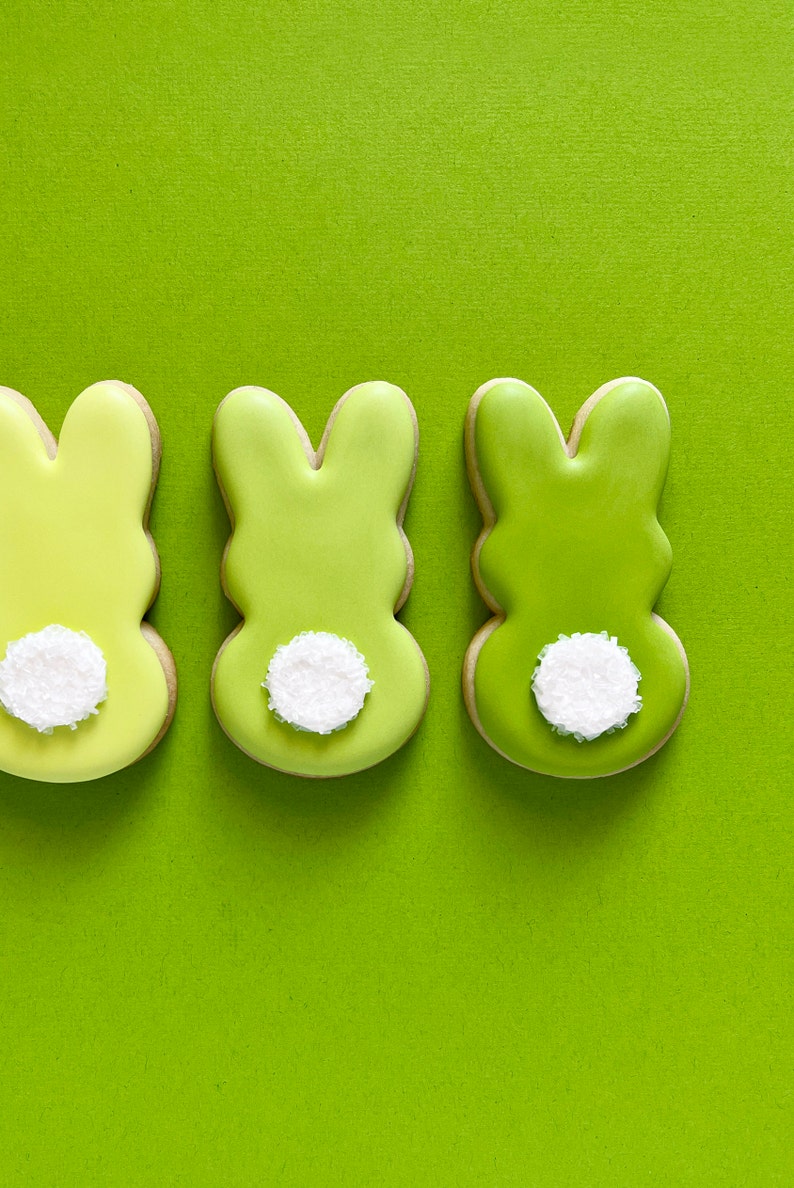 May include: Three green bunny-shaped sugar cookies with white sprinkles on the back. The cookies are arranged in a row on a green background.