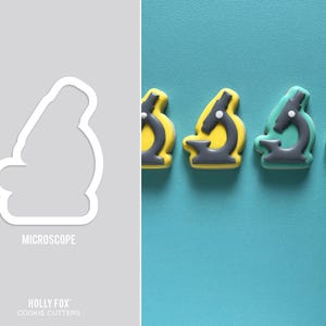 May include: A white microscope-shaped cookie cutter next to several decorated biscuits. The biscuits are yellow and teal with grey and white details. The word "MICROSCOPE" is printed below the cookie cutter.