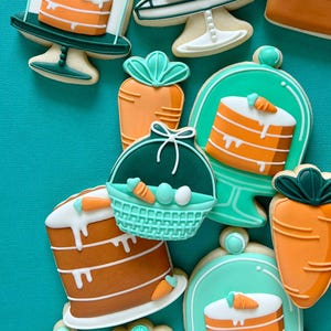 May include: A selection of Easter-themed biscuits. The biscuits are decorated with orange and white icing, featuring carrot and cake designs. Some biscuits are shaped like carrots, cakes, and Easter baskets with eggs. The biscuits are arranged on a teal background.