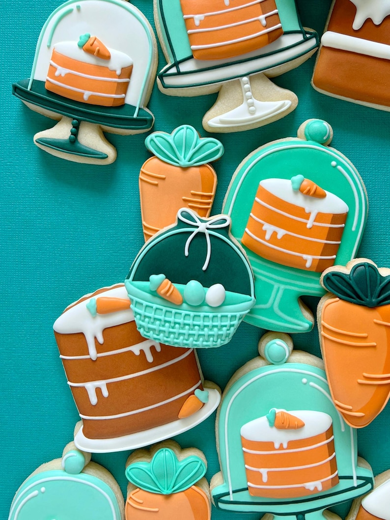 May include: A selection of Easter-themed biscuits. The biscuits are decorated with orange and white icing, featuring carrot and cake designs. Some biscuits are shaped like carrots, cakes, and Easter baskets with eggs. The biscuits are arranged on a teal background.