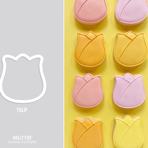 May include: A set of nine cookie cutters shaped like tulips. The cutters are arranged in three rows of three, with each row featuring a different color: pink, yellow, and orange. The cutters are made of metal and have a smooth, polished finish. The text "TULIP" and "HOLLY FOX COOKIE CUTTERS" is visible at the bottom of the image.