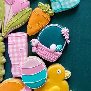 May include: A selection of Easter biscuits, including a yellow duck, a carrot, a basket with eggs, a pink and blue egg, and pink tulips. Other biscuits include a pink and white gingham boot and a bunny face. The biscuits are decorated with colourful icing.