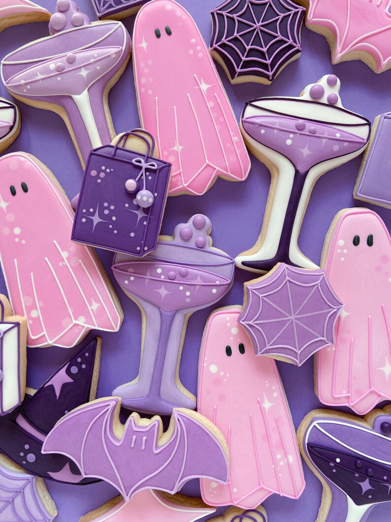 Puede incluir: Galletas de az&uacute;car de Halloween de color morado y rosa decoradas con glaseado. Las galletas tienen forma de fantasmas, murci&eacute;lagos, telara&ntilde;as, sombreros de bruja, copas de champ&aacute;n y bolsas de la compra.