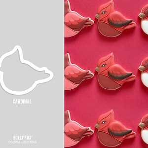 May include: A white cardinal-shaped cookie cutter with the text CARDINAL and HOLLY FOX COOKIE CUTTERS printed below it, alongside a grid of decorated cardinal cookies featuring red, dark red, and white icing on a solid red background.
