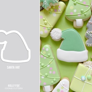 May include: A white cookie cutter in the shape of a Santa hat. The cookie cutter is on a gray background. The text "SANTA HAT" is below the cookie cutter. The text "HOLLY FOX" and "COOKIE CUTTERS" is below the text "SANTA HAT".