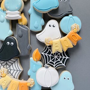 May include: A collection of Halloween-themed sugar cookies decorated with various designs, including ghosts, pumpkins, candles, and spiderwebs. The cookies are decorated in shades of blue, white, orange, and gray.