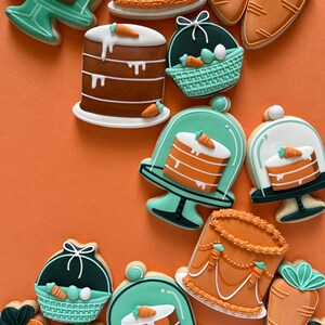 May include: A selection of Easter biscuits in the shapes of cakes, carrots, and baskets. The biscuits are decorated with orange, brown, white, and teal icing. They are arranged on an orange background.