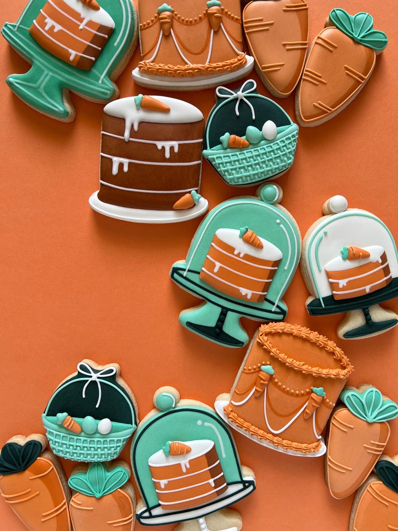 May include: A selection of Easter biscuits in the shapes of cakes, carrots, and baskets. The biscuits are decorated with orange, brown, white, and teal icing. They are arranged on an orange background.