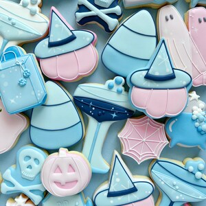 Puede incluir: Una colecci&oacute;n de galletas de az&uacute;car con tem&aacute;tica de Halloween decoradas con glaseado azul, rosa y blanco. Las galletas presentan varios dise&ntilde;os, incluyendo fantasmas, calabazas, sombreros de bruja, ma&iacute;z dulce y una calavera y huesos cruzados.