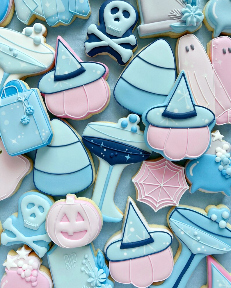 Puede incluir: Una colecci&oacute;n de galletas de az&uacute;car con tem&aacute;tica de Halloween decoradas con glaseado azul, rosa y blanco. Las galletas presentan varios dise&ntilde;os, incluyendo fantasmas, calabazas, sombreros de bruja, ma&iacute;z dulce y una calavera y huesos cruzados.
